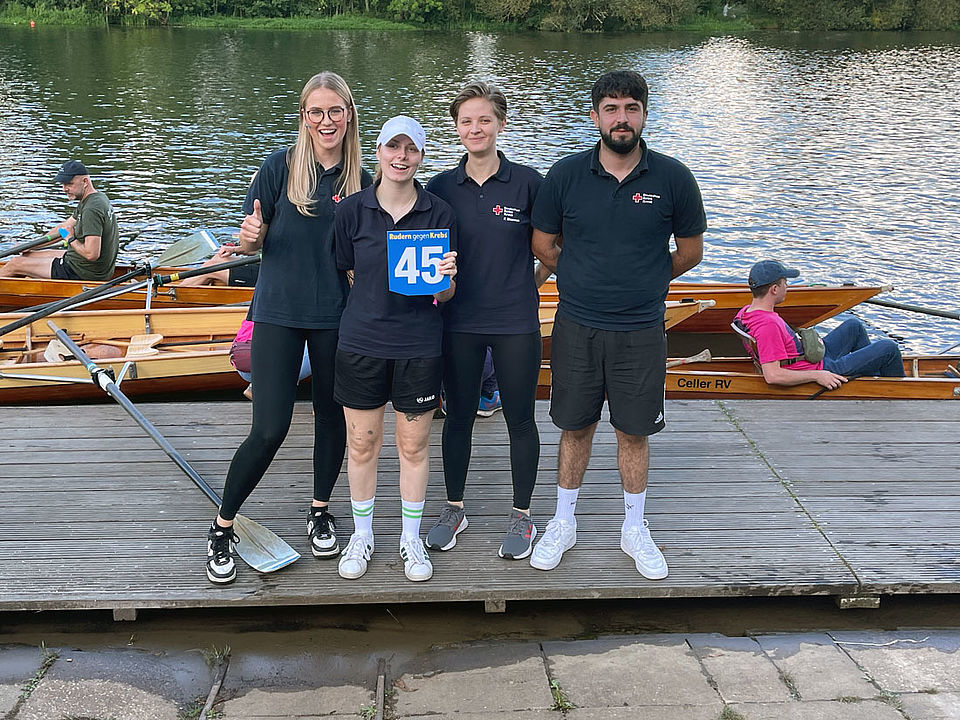 Vier Kolleg*innen freuen sich über den ersten Platz. Personen stehen auf dem Bootssteg nebeneinander, nach dem Aussteigen aus dem Ruderboot.