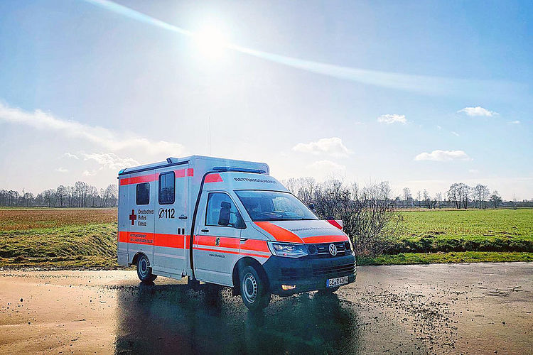 Ein Mehrzweckfahrzeug (MZF) des Rettungsdienstes steht vor einem sonnenbeschienenen Feld. Die Szene wirkt ruhig und ländlich.