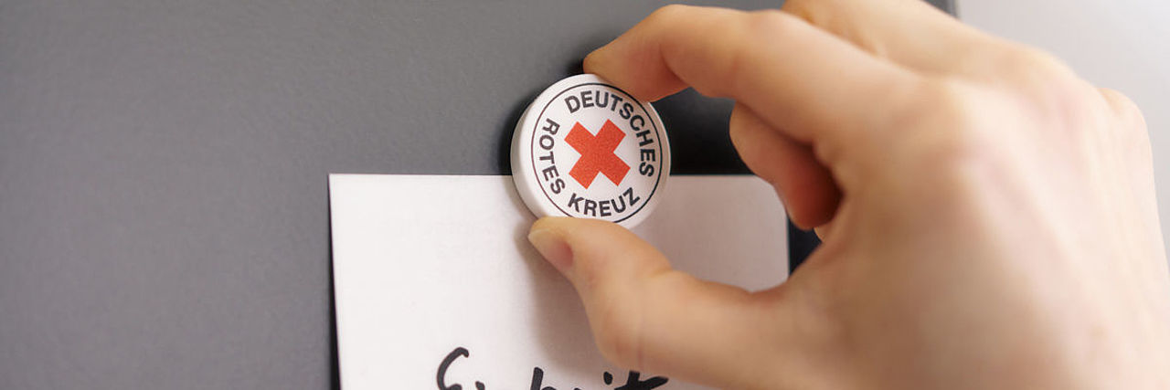 Symbolfoto Magnet mit DRK-Logo befestigt Zettel mit dem Schriftzug Grundsatz ‚Einheit‘ auf einer Tafel