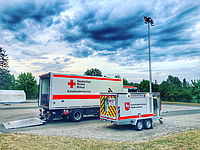 Fahrzeuge des Katastrophenschutzes, darunter ein Lastwagen und ein Anhänger mit Stromaggregat und Lichtmast, stehen unter einem teils blauen Himmel mit dunklen Wolken.