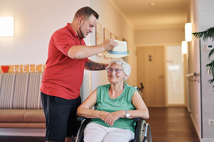 Mitarbeiter setzt einer Seniorin einen Hut auf, um sie mit der Kopfbedeckung vor Sonneneinstrahlung zu schützen