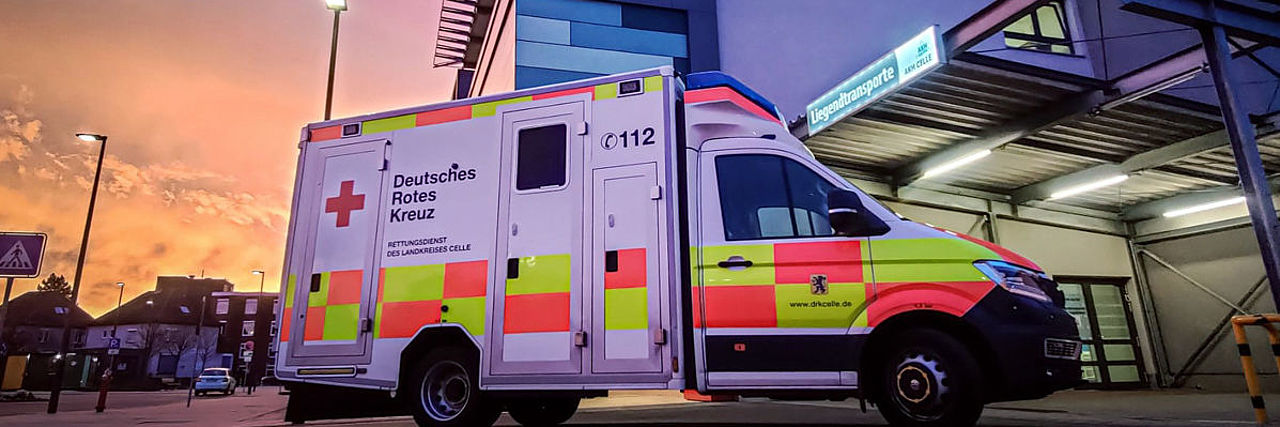 Ein Rettungswagen steht schräg von der Seite vor dem Gebäude des AKH in Celle. Die Szene ist in warmes Licht des Sonnenaufgangs getaucht.