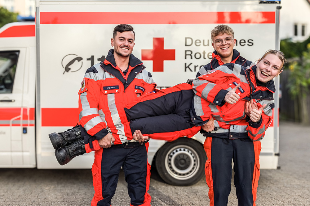 Drei Bundesfreiwillige in Einsatzbekleidung stehen vor einem Rettungswagen. Die Kollegin in der Mitte wird von ihren beiden männlichen Kollegen auf Händen getragen – alle lächeln.